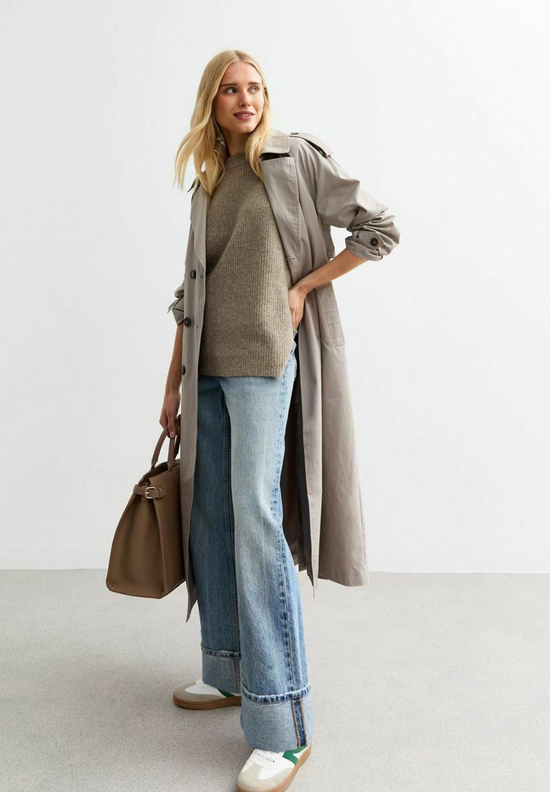 Ljusgrå trenchcoat över en grön ribbad tröja, parat med vida blå jeans och vita sneakers. Håller en brun handväska.