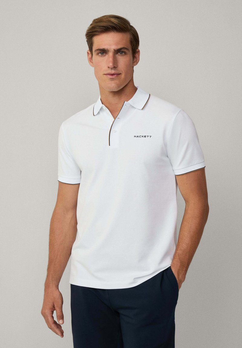 Polo bianco a maniche corte, con colletto strutturato e accentato in nero attorno al colletto e alle maniche. Presenta la parola "HACKETT" stampata in nero.