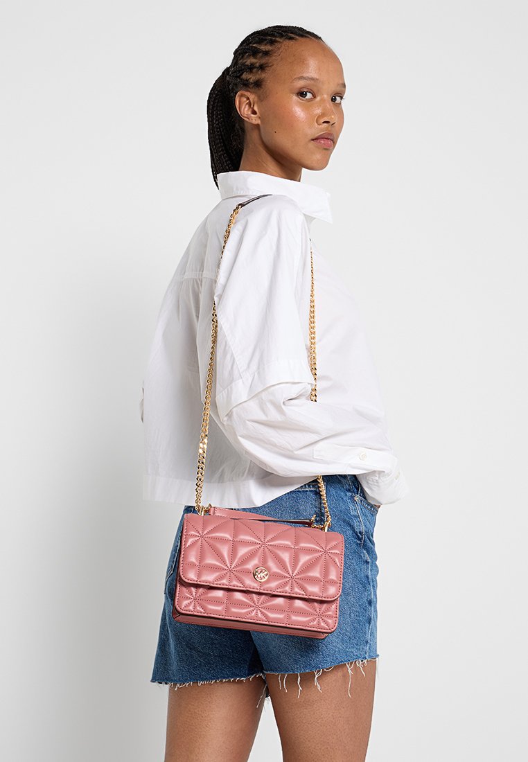 Sac en cuir matelassé rose avec une bandoulière en chaîne dorée, doté d'un rabat à l'avant et d'un accent de logo circulaire. Positionné à côté d'un mannequin.