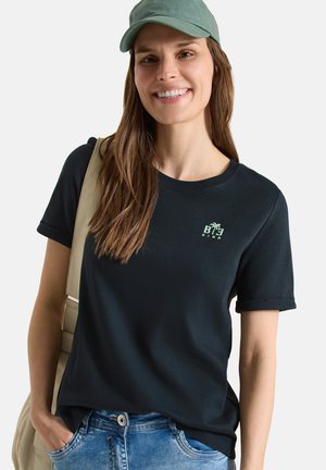 Vrouw draagt een zwart "Be Kind" t-shirt, groene pet, beige rugzak en blauwe spijkerbroek, glimlachend tegen een effen achtergrond.