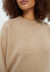 Beidzas crewneck džemperis ar mīkstu tekstūru, raglāna piedurknēm un subtilu rakstu audumā. Tuvplāns skats uz kakla un plecu zonu.