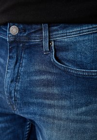 Primer plano de la cintura y el bolsillo de unos jeans de mezclilla azul, mostrando un botón metálico y una trabilla para el cinturón, usados con una camiseta negra.