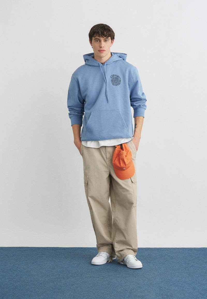 Joven con sudadera azul con capucha, pantalones cargo beige, zapatillas blancas, sosteniendo una gorra naranja a su lado contra un fondo blanco liso.