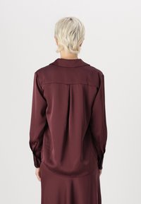 Bordová saténová blúzka s dlhými rukávmi, golierom, detailom bočného sklzu na chrbte a zapínaním na gombíky na rukávoch. Hladká textúra a jemne zakrivený spodný lem.