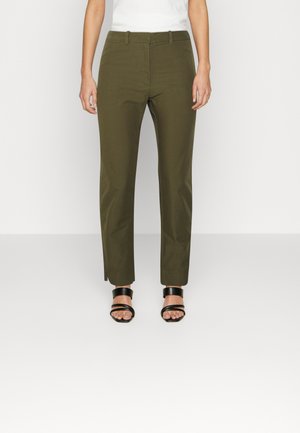 Kvinde iført olivengrønne skræddersyede bukser og sorte åbne tåhøjhælede sandaler, stående foran en helt hvid baggrund.