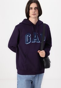 Felpa viola con tasca frontale e grafica "GAP" in blu scuro. Abbinata a jeans azzurri e una piccola borsa a tracolla nera.