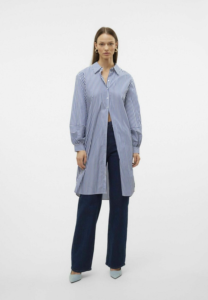 Camicia a righe con bottoni, maniche lunghe, blu e bianco, vestibilità oversize, indossata sopra jeans a zampa d'elefante blu scuro, abbinata a tacchi azzurri.