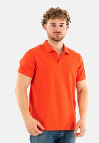 Polo naranja, de manga corta, con cuello, mezcla de algodón, presenta un pequeño logo azul en el pecho, combinado con vaqueros azules.