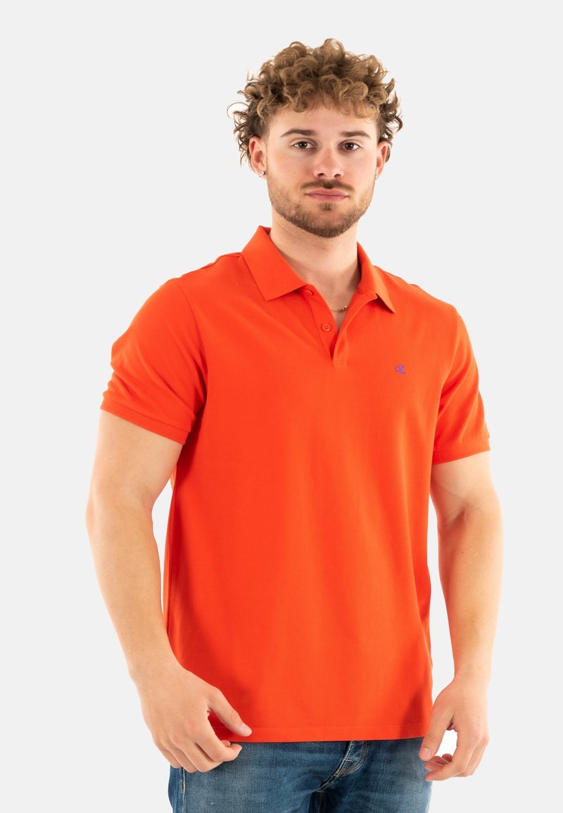 Polo naranja, de manga corta, con cuello, mezcla de algodón, presenta un pequeño logo azul en el pecho, combinado con vaqueros azules.