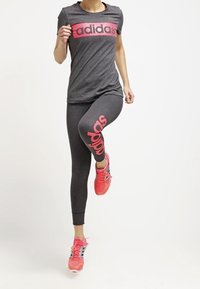 Person i mörkgrå Adidas-outfit med rosa logotyp, som lyfter ena knät, iförd klarröda löparskor, på vit bakgrund.