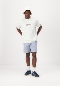 Camiseta de algodón verde claro con gráfico púrpura, pantalones cortos a rayas azules, sombrero blanco tipo cubo, zapatillas navy con rayas azul claro, calcetines blancos.