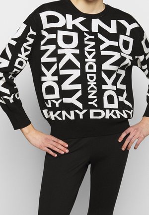 Zwarte trui met grote witte herhaalde DKNY-logo's gedragen door een persoon met handen in de heupen, gecombineerd met een zwarte broek.