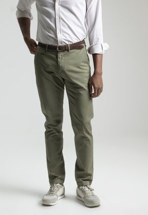 Mężczyzna ubrany w białą podwiniętą koszulę, oliwkowozielone spodnie z brązowym paskiem oraz jasnoszare tenisówki, stojący na jednolitym tle.