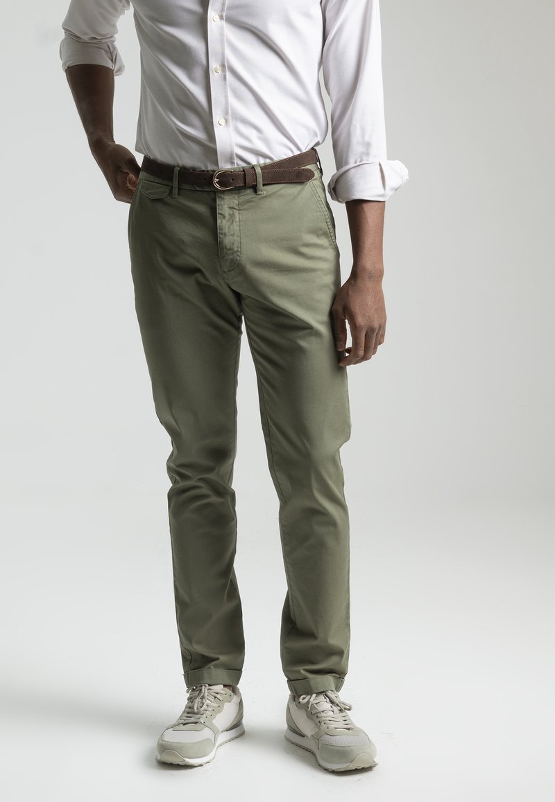 Homme portant une chemise blanche retroussée, un pantalon vert olive avec une ceinture marron, et des baskets gris clair, debout devant un fond uni.