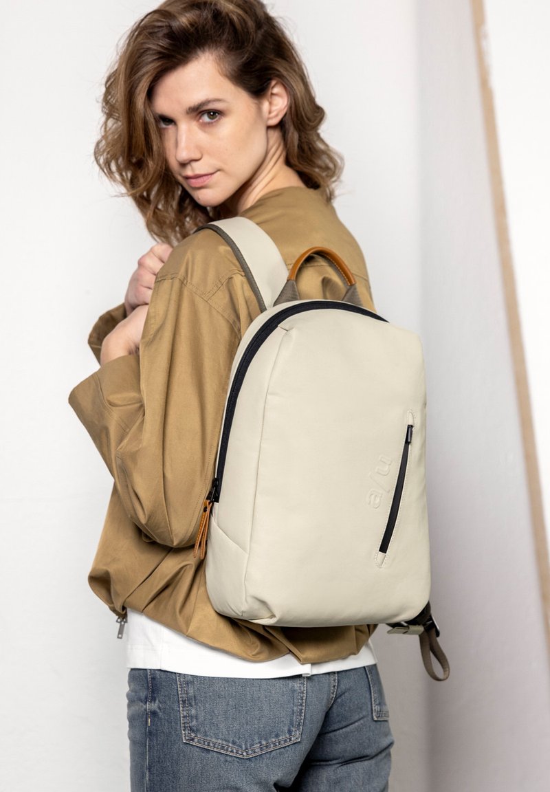 Beige rugzak met een gladde textuur, bovenhandgreep en een contrasterende donkere rits, gedragen over een bruin shirt en lichte jeans tegen een neutrale achtergrond.