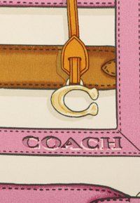 Leren band met oranje accent, goudkleurige hardware met een "C" logo, tegen een roze en witte patroon achtergrond met "COACH" merknaam.