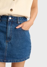 Gonna di jeans di colore blu scuro con un design pulito, tasca frontale e dettagli di cucitura visibili. Il materiale appare casual e texturizzato.