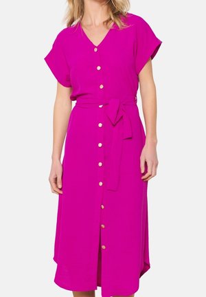 Femme portant une robe rose vif boutonnée devant, manches courtes et ceinture nouée à la taille sur fond blanc.