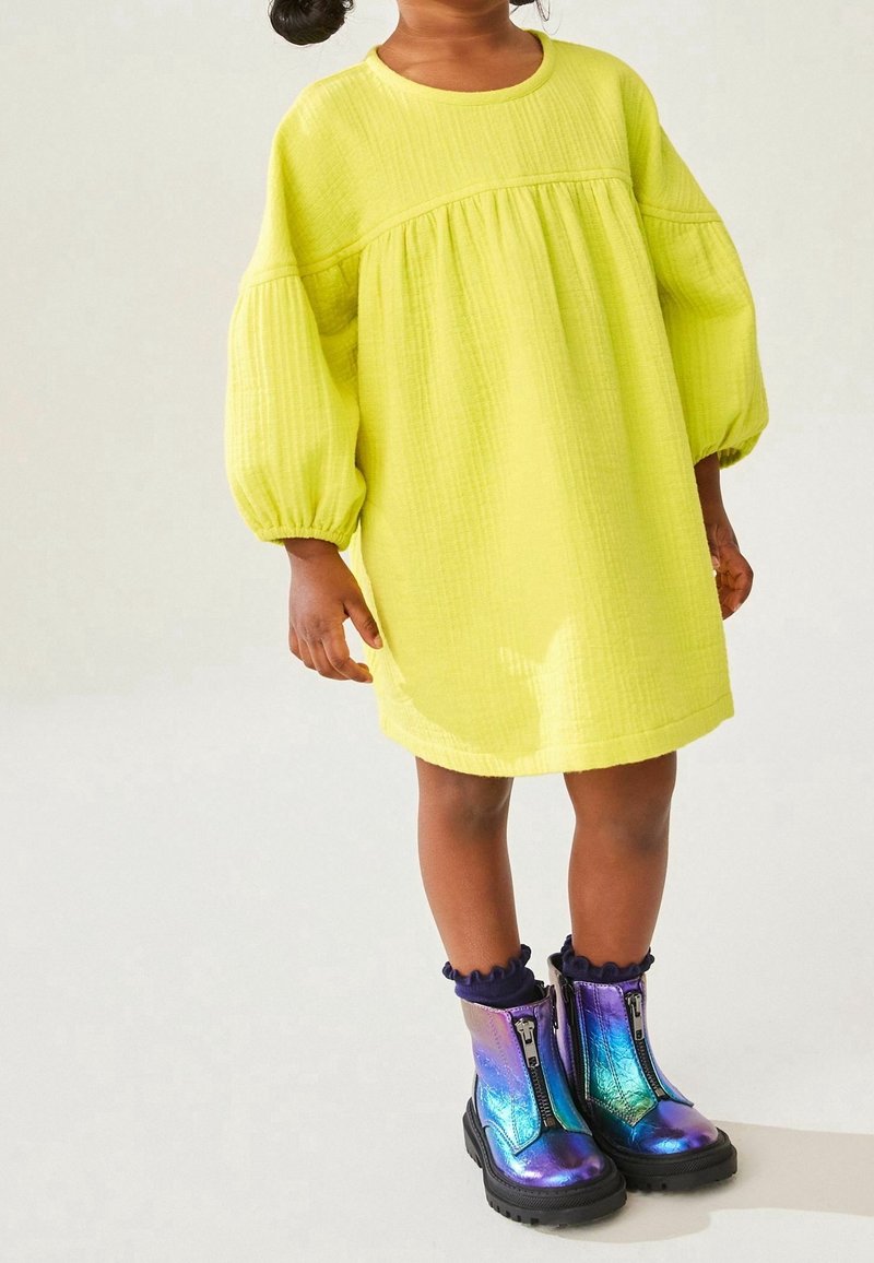 Enfant portant une robe jaune ample à manches bouffantes et des bottes à fermeture éclair devant brillantes et iridescentes avec des semelles noires.