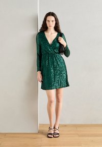 Abito verde con paillettes, maniche lunghe, scollatura a V, vita cintrata e gonna svasata. Abbinato a sandali neri con cinturini e una borsa piccola.