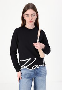 Čierna mikina s bielym logom "Karl", okrúhly výstrih, nosená s svetlo modrými džínsami. Doplnkom je svetlo béžová taška s popruhom "HUGO".