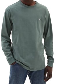 Mann trägt ein grünes Langarmshirt von Vans mit blauen Jeans, eine Hand in der Tasche und die andere entspannt an seiner Seite.