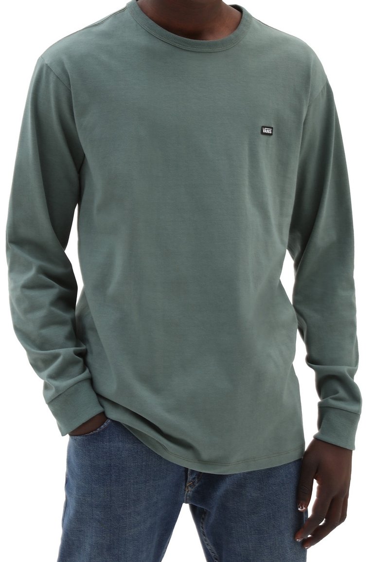 Mann trägt ein grünes Langarmshirt von Vans mit blauen Jeans, eine Hand in der Tasche und die andere entspannt an seiner Seite.