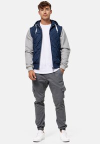 Chaleco acolchado navy con mangas y capucha grises, usado sobre una camiseta blanca; combinado con pantalones cargo grises y zapatillas blancas.