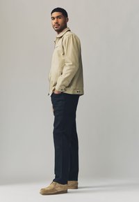 Beige lærredsjakke, sorte bukser, brune sko. Jakken har lynlås- og knaplukning, sidelommer og en krave. Glat tekstur over det hele.