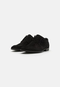 Chaussures lacées en suede noir avec une finition lisse, un design à bout pointu et des détails de couture minimaux. Semelle en caoutchouc plate et talon subtil.