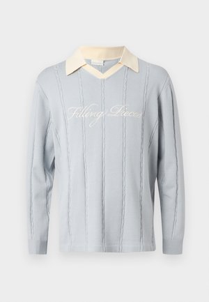 Lys blå strikke genser med vertikale kabelmønstre, kremfarget V-hals og kursiv "Filling Pieces"-tekst over brystet.