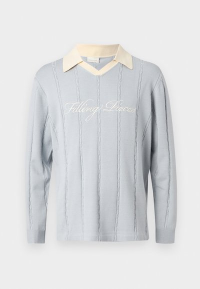 Ανοιχτό γαλάζιο πλεκτό πουλόβερ με κάθετα σχέδια καλωδίων, γιακά σε σχήμα V σε εκρού χρώμα και καλλιγραφικό κείμενο "Filling Pieces" σε όλο το στήθος.