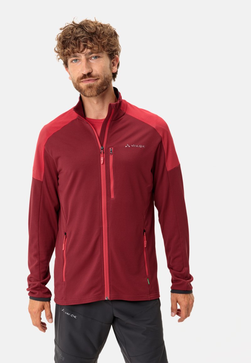 Giacca rossa con zip, maniche in un rosso scuro a contrasto, due tasche anteriori con zip e tessuto strutturato. Indossata sopra una maglietta rossa coordinata.