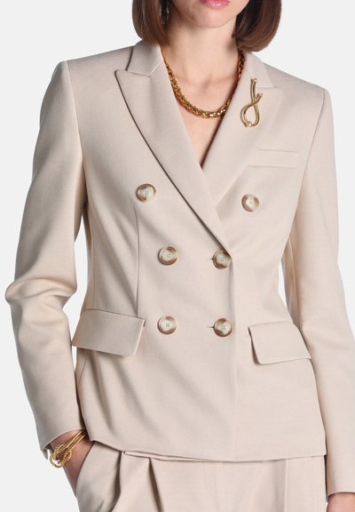 Femme portant un blazer beige croisé à six boutons, collier chaîne en or, broche en or sur le revers et bracelet en or, main dans la poche.