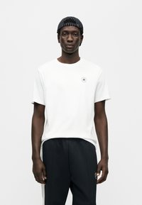 Λευκό κοντομάνικο μπλουζάκι με στρογγυλή λαιμόκοψη· διαθέτει κυκλική λογότυπο σε μπλε και κόκκινο στο αριστερό στήθος. Μαύρο παντελόνι με κάθετη λεπτομέρεια ρίγας.