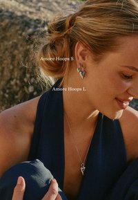 Silver hoop earrings with a geometric design. One pair is larger than the other, both showcasing a polished finish. Neckline features a heart pendant.