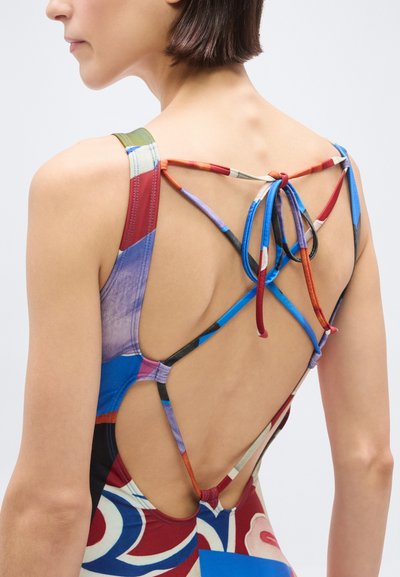 Γυναίκα που φοράει πολύχρωμο ολόσωμο μαγιό με ανοιχτή πλάτη και λεπτές, διασταυρωμένες τιράντες που δένονται σε φιόγκο.