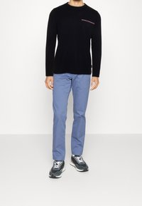 Schwarzes langärmliges Shirt mit kleinem Brustdruck, kombiniert mit hellblauen Hosen und dunklen Sneakers mit einer weißen Sohle.