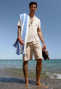 Homme debout pieds nus dans l'eau peu profonde de la mer, portant un short beige et une chemise, tenant des sandales et une serviette rayée bleue et blanche sur l'épaule.