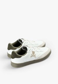 Zapatillas blancas de corte bajo con suelas y tacones verde oliva, diseño con cordones y un logo de calavera y tibias en color beige en el lateral.