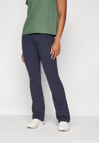 Sötétkék, széles szárú leggings sima felülettel, laza zöld pólóval párosítva. Fehér sportcipők lila kiegészítőkkel teszik teljessé az öltözetet.