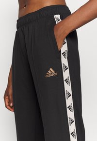 Černé sportovní kalhoty z měkké látky s elastickým pasem, bočními kapsami a bílou stuhou s logem Adidas podél nohou.
