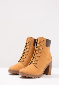 Bottes basses en cuir beige avec un talon haut, des lacets jaunes et un col en cuir marron. Elles présentent une semelle en caoutchouc texturée et des détails de marque.