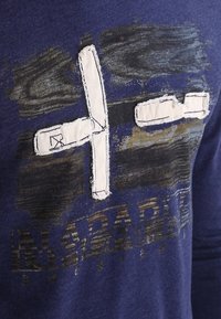 Tmavě modrá košile s dlouhým rukávem, která má texturovanou grafiku s bílým křížovým designem, doplněnou o švy a opotřebované barevné variace.