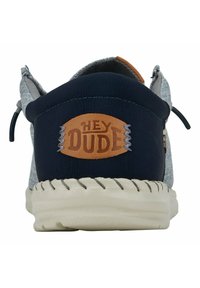 Scarpa in tessuto grigio con tacco blu navy, caratterizzata da un'etichetta in pelle marrone con la scritta "HEY DUDE" in rilievo. Suola spessa di colore crema con cuciture a contrasto.