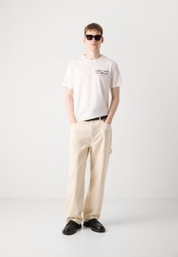 Hvid t-shirt med rød og blå grafisk print, beige brede bukser og sorte solbriller. Sorte sko fuldender outfittet.