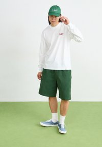 Witte blouse met lange mouwen en een rood logo, groene shorts, groene pet en blauwe geruite sneakers, tegen een lichte achtergrond en een groene vloer.
