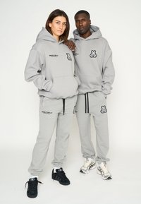 Las sudaderas y pantalones de chándal grises presentan logotipos bordados, con un ajuste relajado y cordones. Los modelos llevan zapatillas negras y plateadas.