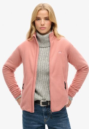 Blonde Frau trägt eine rosafarbene Reißverschluss-Fleecejacke über einem grauen Strickrollkragenpullover und bluejeans mit einem dekorativen Gürtel.