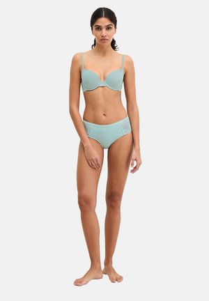 Woman standing barefoot wearing a light blue bra with adjustable straps and matching high-cut underwear with lace side details.